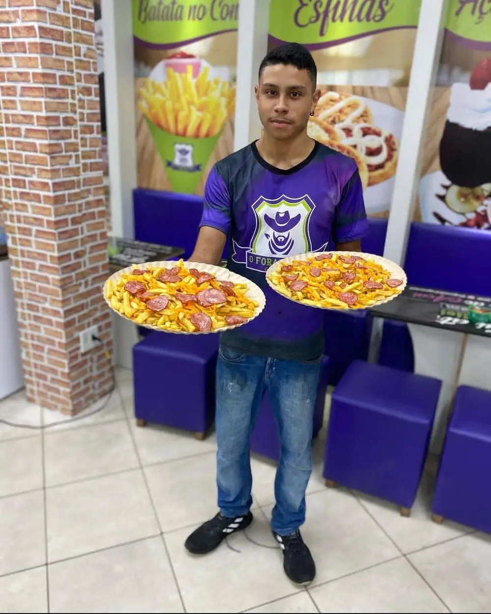 Funcionário de uniforme roxo segurando duas travessas de batata com calabresa, na frente dos banners de batata no cone e esfihas